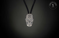 Silver skull pendant intricately carved with an ornate pattern and blue gem eyes, on a black braided cord.
