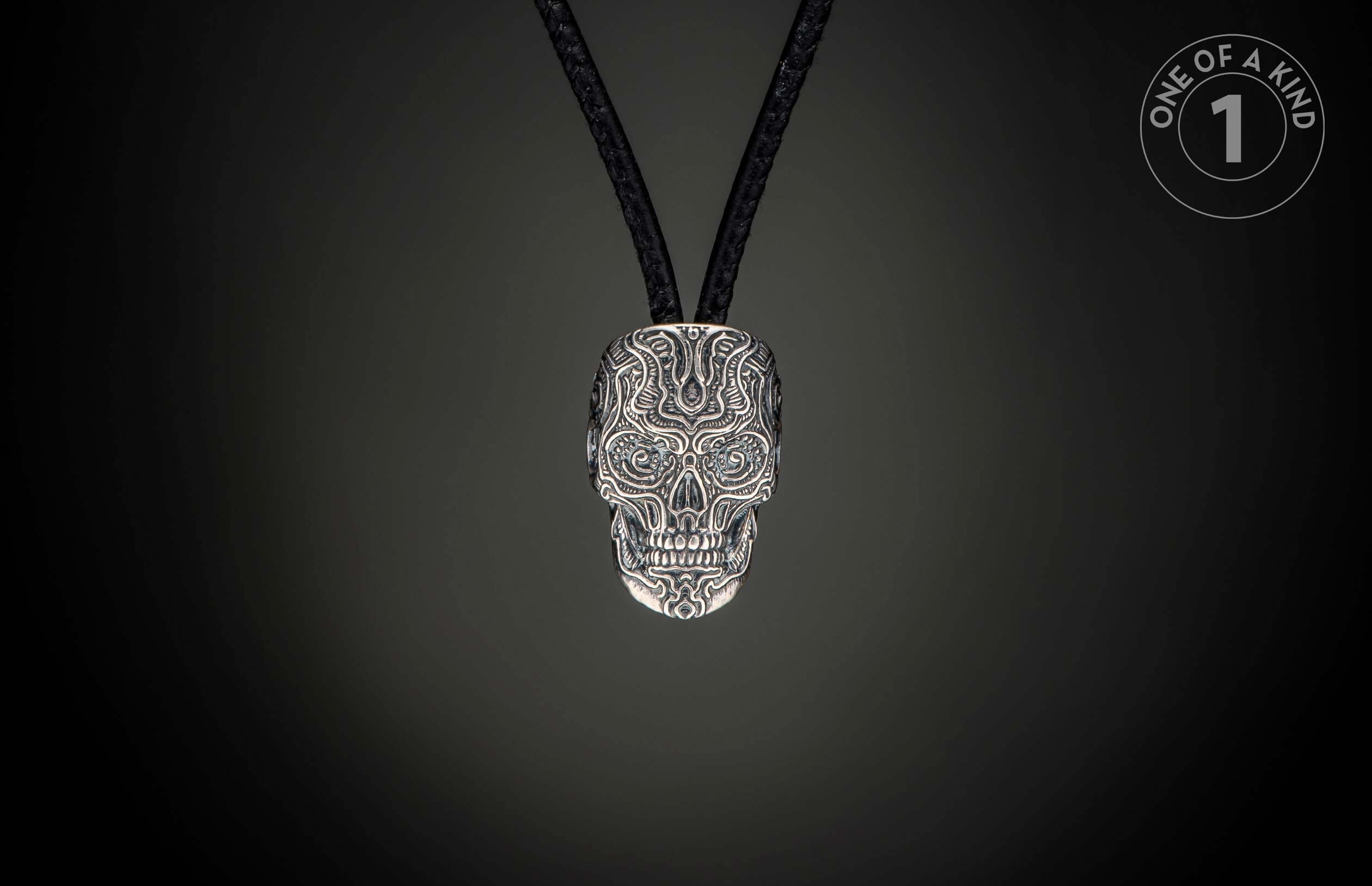 Silver skull pendant intricately carved with an ornate pattern and blue gem eyes, on a black braided cord.