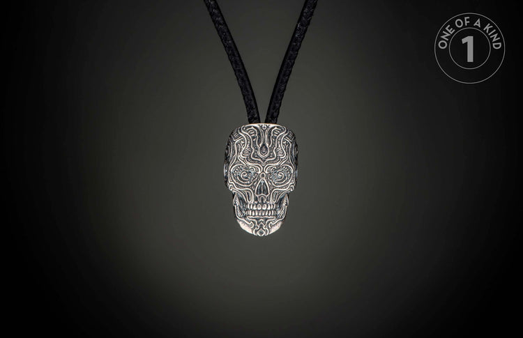 Silver skull pendant intricately carved with an ornate pattern and blue gem eyes, on a black braided cord.