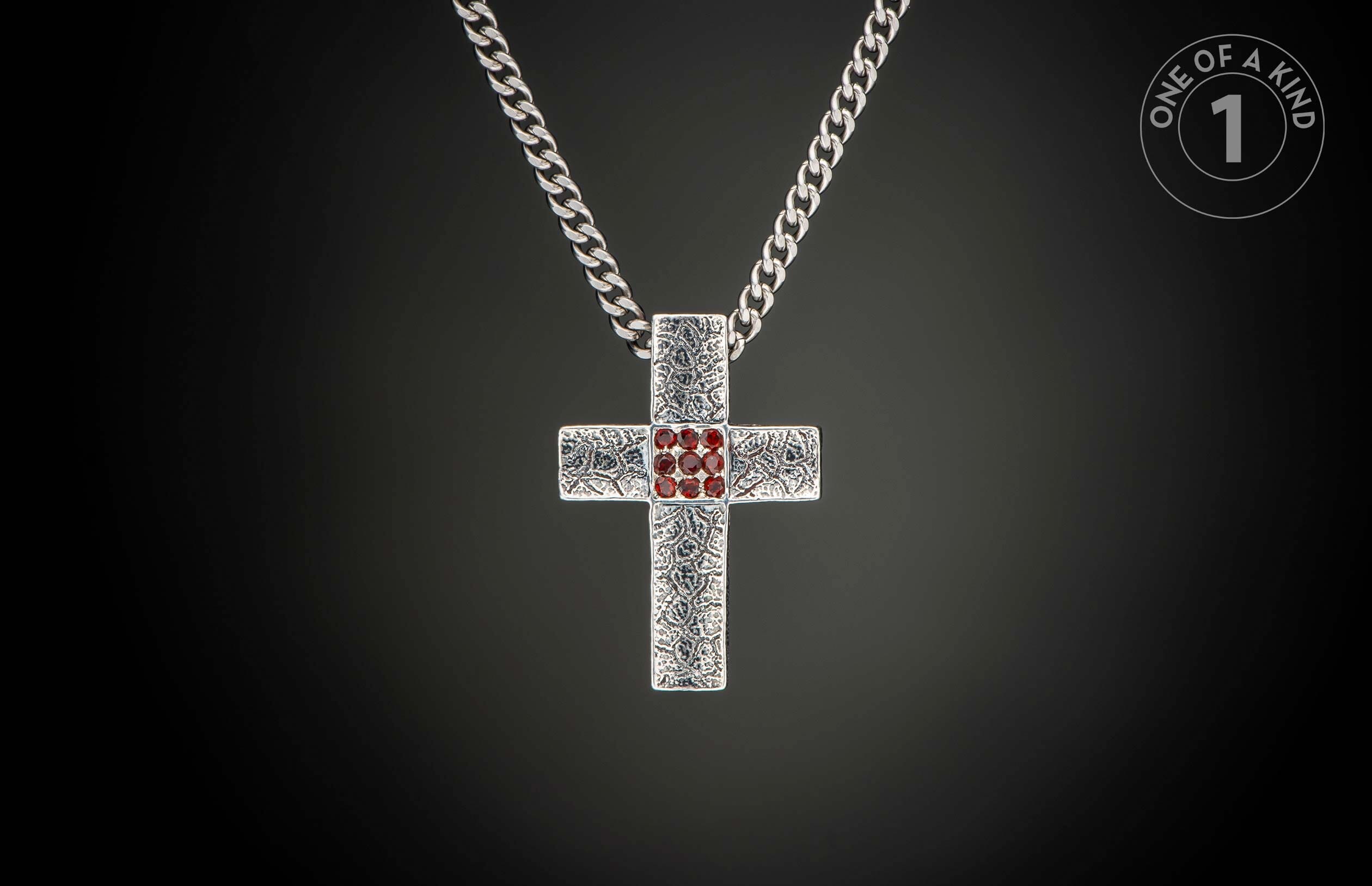 Silver cross pendant with a textured pattern and red gemstones in the center, hanging from a curb chain.