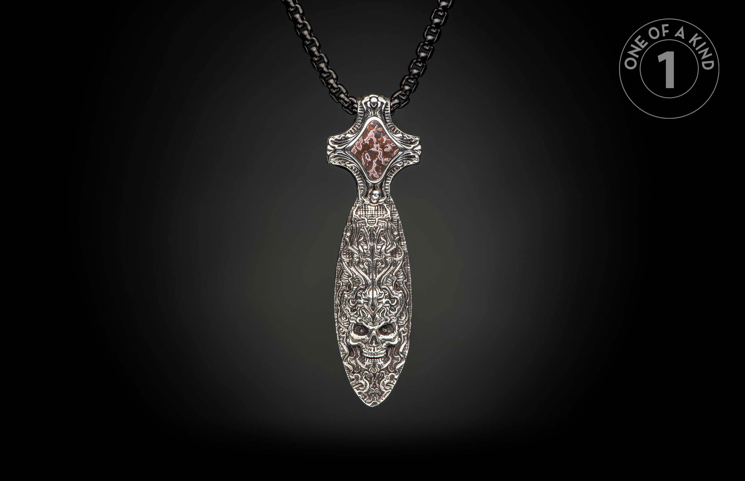 Ornate silver dagger pendant with skull and intricate carvings, featuring a reddish-brown stone inlay, on a dark chain.