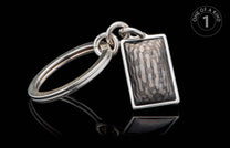 Silver-toned metal keychain with a rectangular charm featuring a mottled gray irregular pattern, reflecting on a black surface.