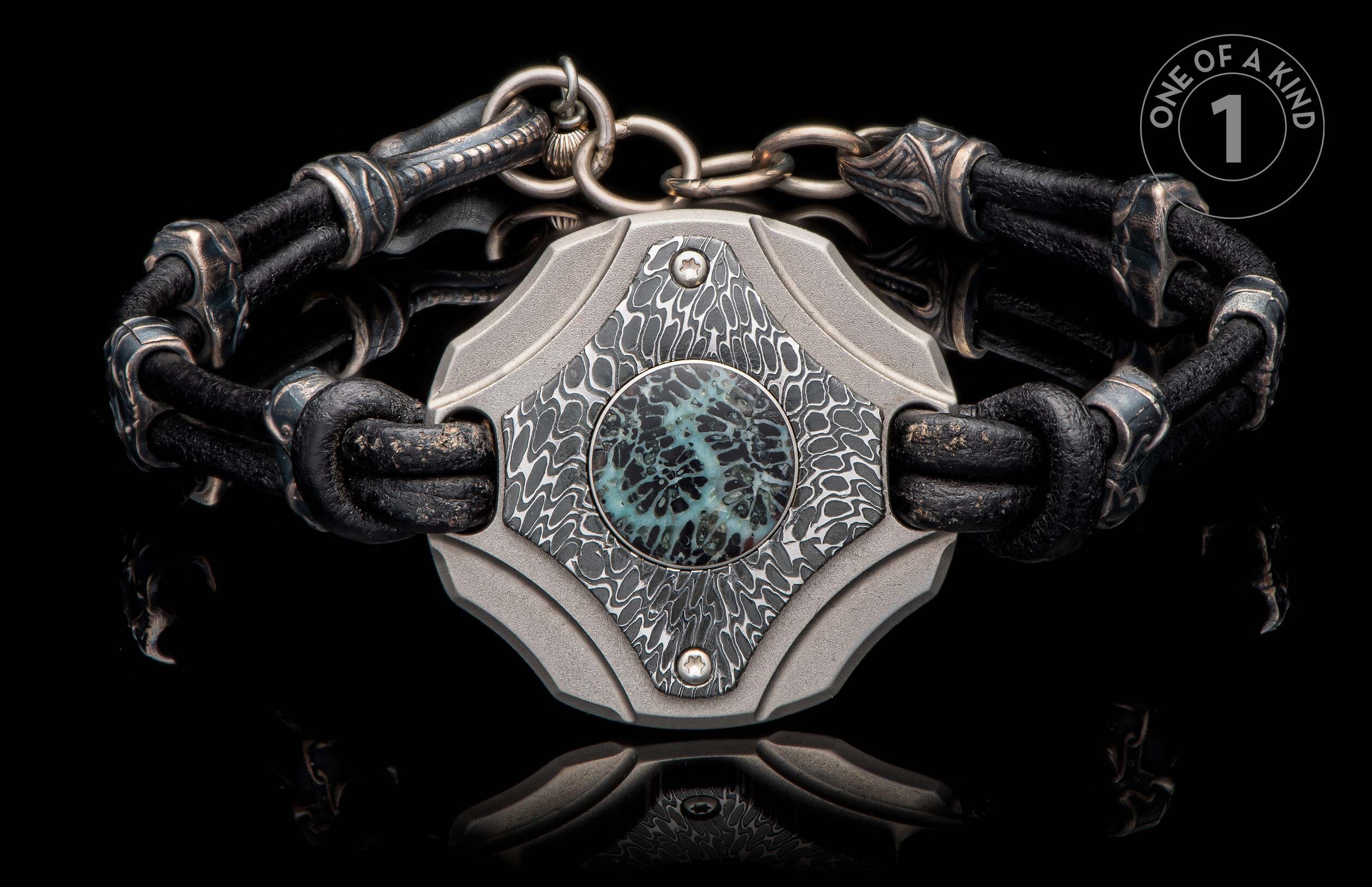 Black leather bracelet with textured metal links and an intricate patterned centerpiece with a blue-green round inlay.