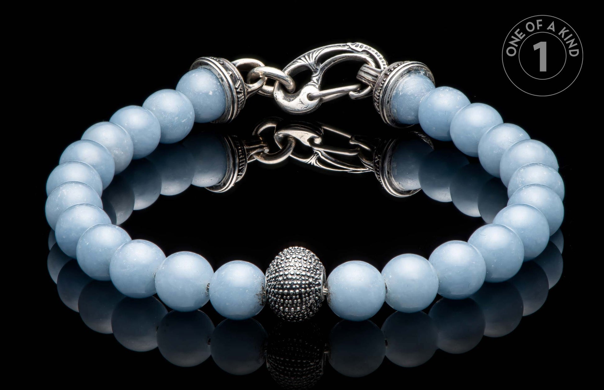 Light blue beaded bracelet featuring a textured silver-tone centerpiece bead and an intricate silver-tone toggle clasp, reflected on black.