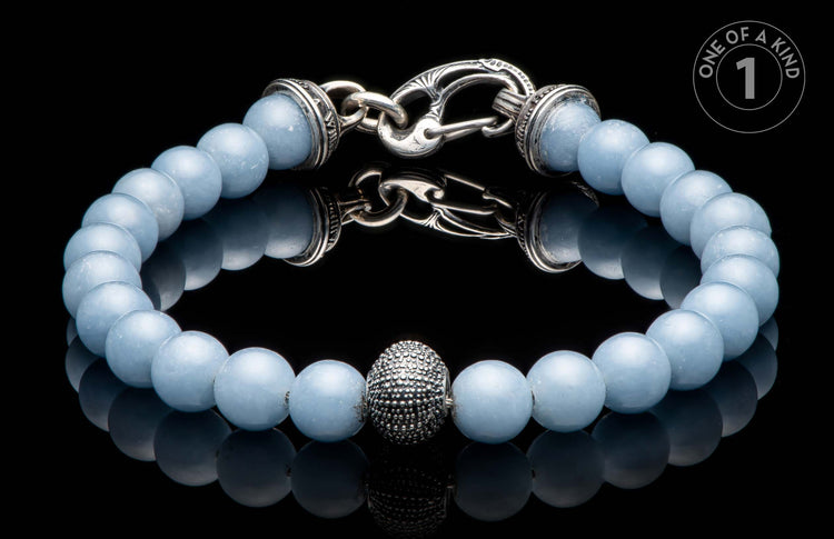 Light blue beaded bracelet featuring a textured silver-tone centerpiece bead and an intricate silver-tone toggle clasp, reflected on black.