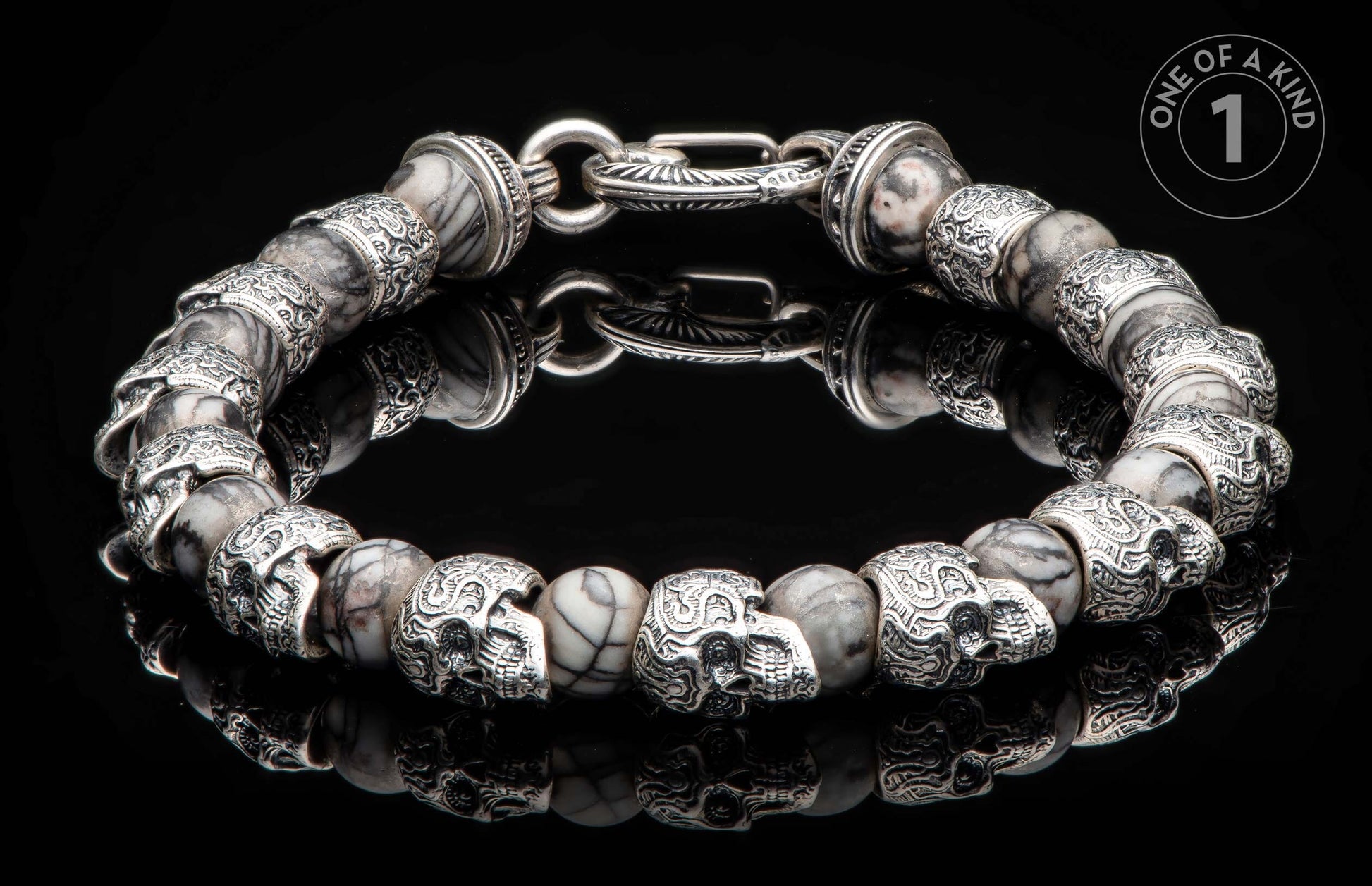 Silver-tone and marbled stone bracelet with skull beads and an ornate clasp, lying on a reflective black surface.