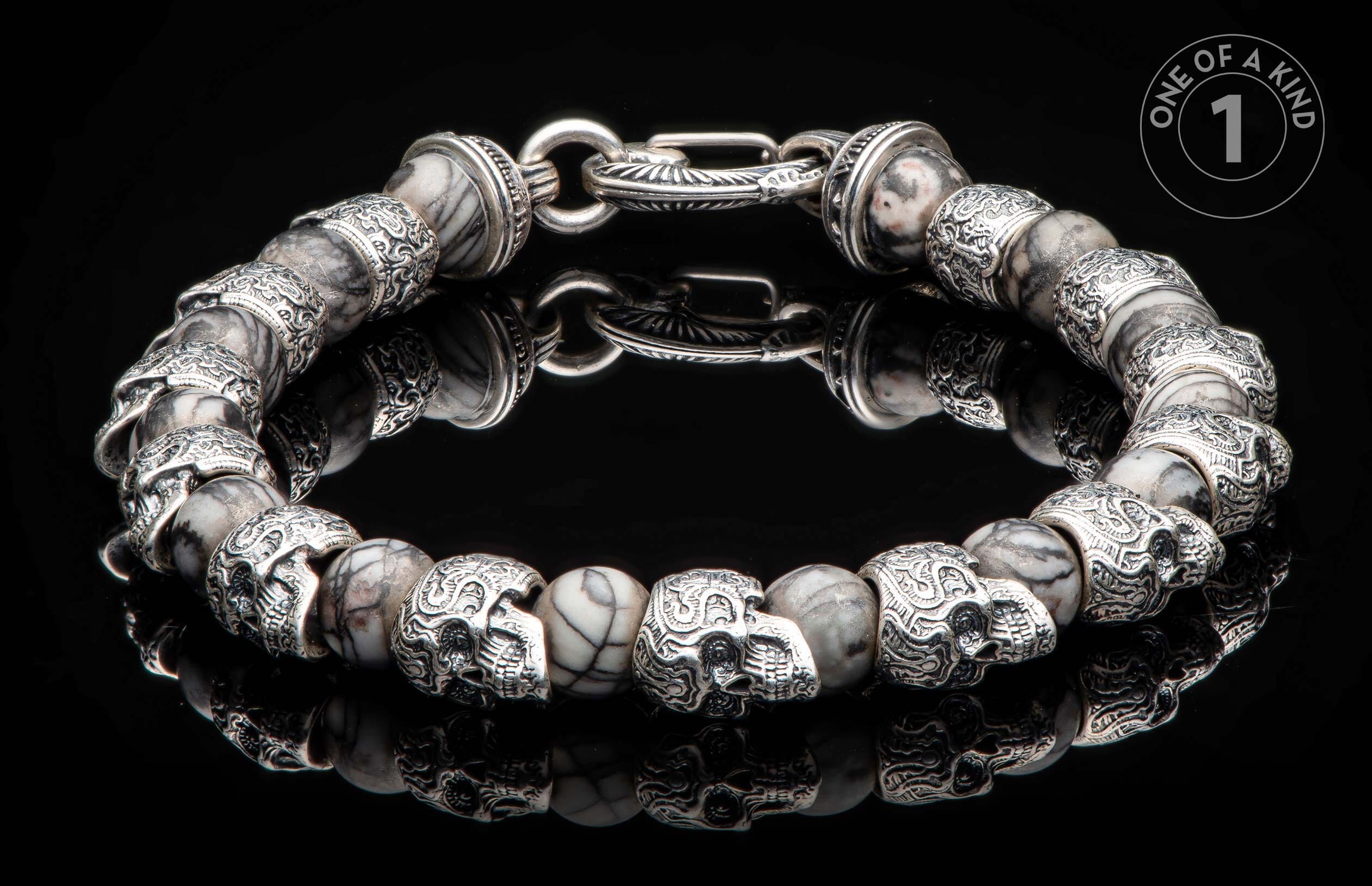 Silver-tone and marbled stone bracelet with skull beads and an ornate clasp, lying on a reflective black surface.