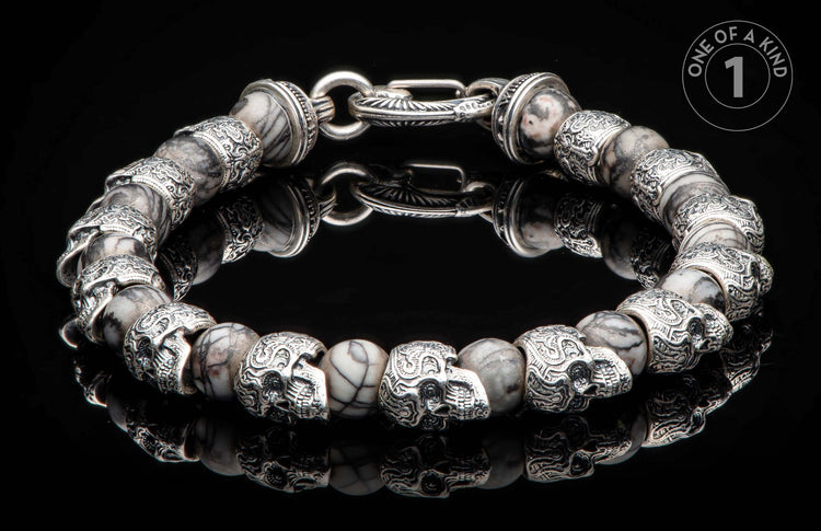 Silver-tone and marbled stone bracelet with skull beads and an ornate clasp, lying on a reflective black surface.
