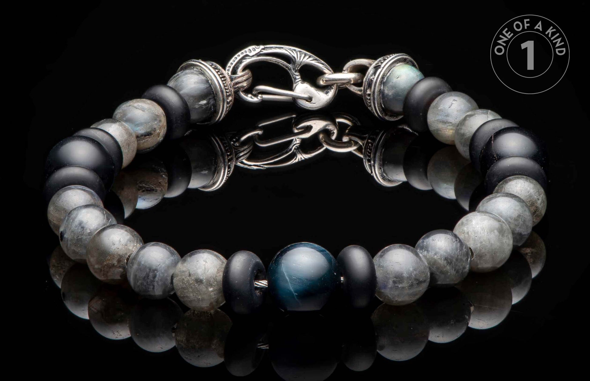 Beaded bracelet with black, grey, and blue stone beads and an ornate silver clasp, reflecting on a dark surface.
