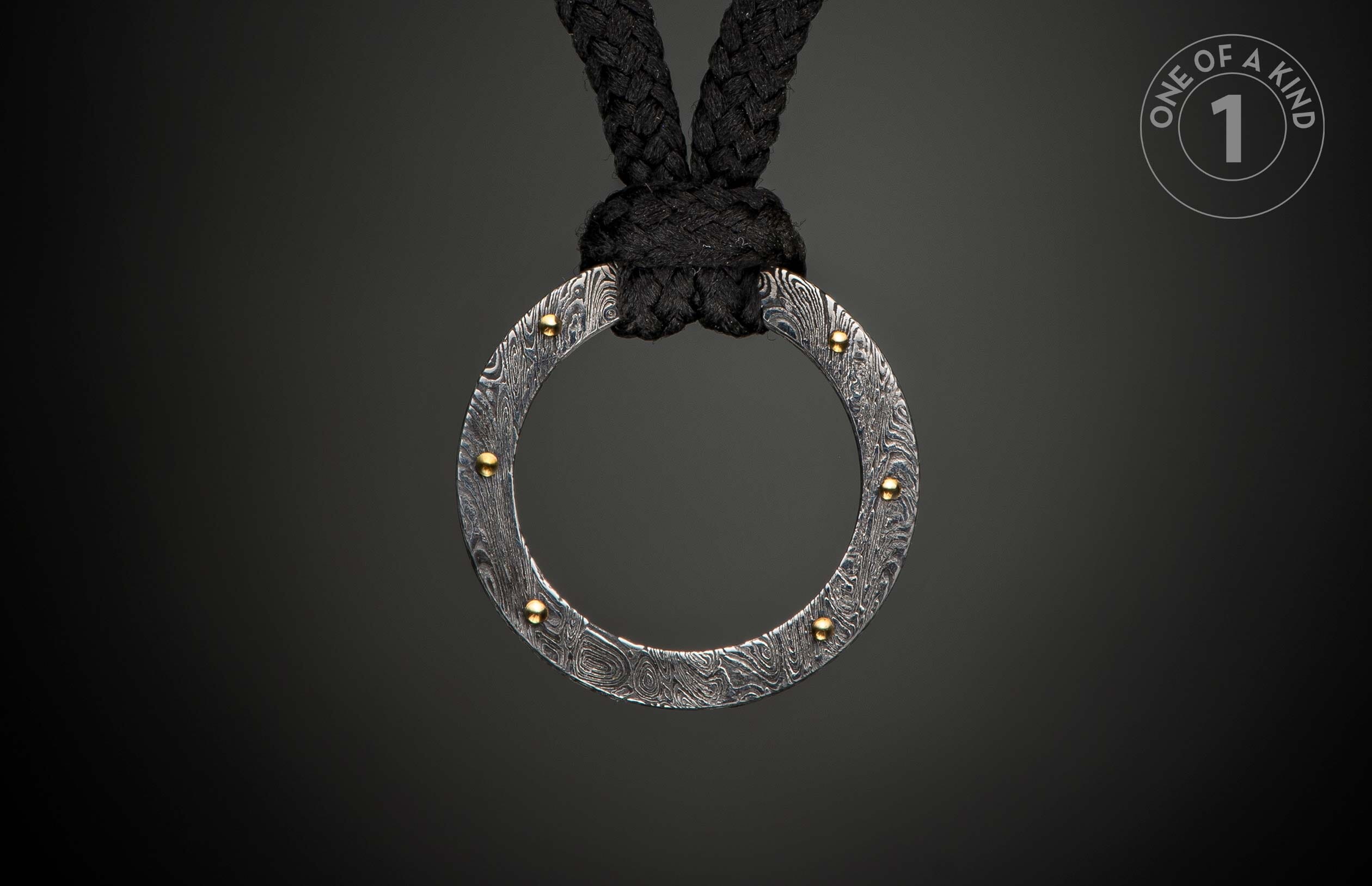 Damascus steel ring pendant with swirling patterns and gold rivets, suspended from a black braided rope.