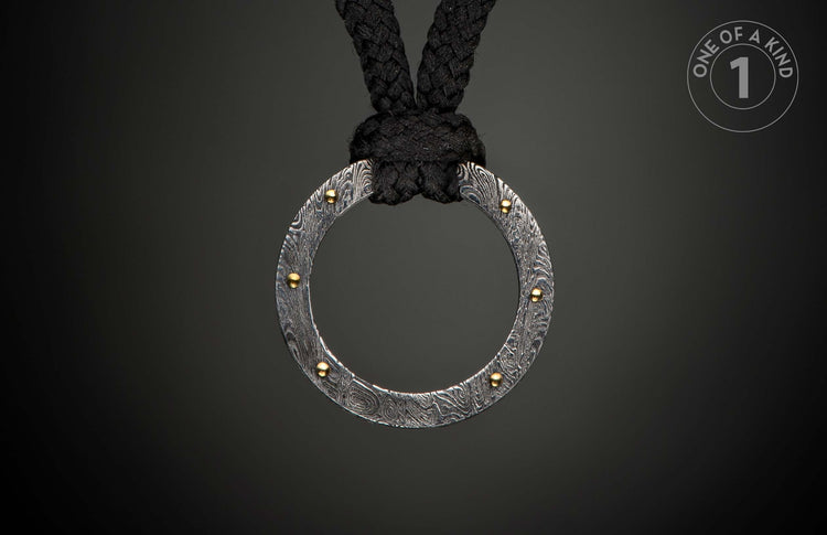 Damascus steel ring pendant with swirling patterns and gold rivets, suspended from a black braided rope.