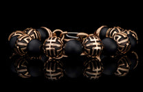 Black beaded bracelet with ornate gold-tone patterned beads and a metallic clasp, reflected on a dark surface.