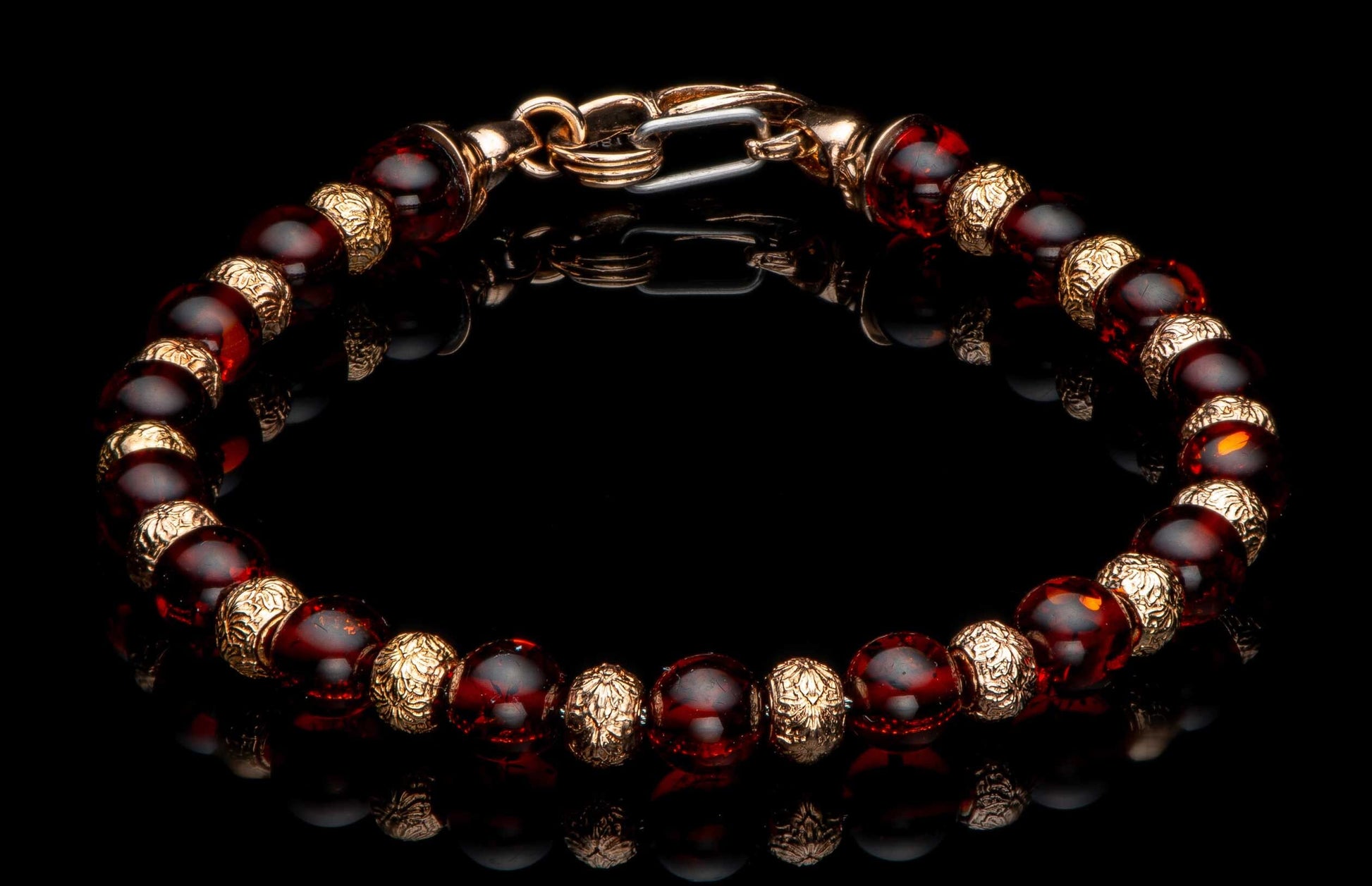 Beaded bracelet with reddish-brown amber-like beads, intricately patterned gold-toned spacers, and a lobster clasp on a reflective black surface.