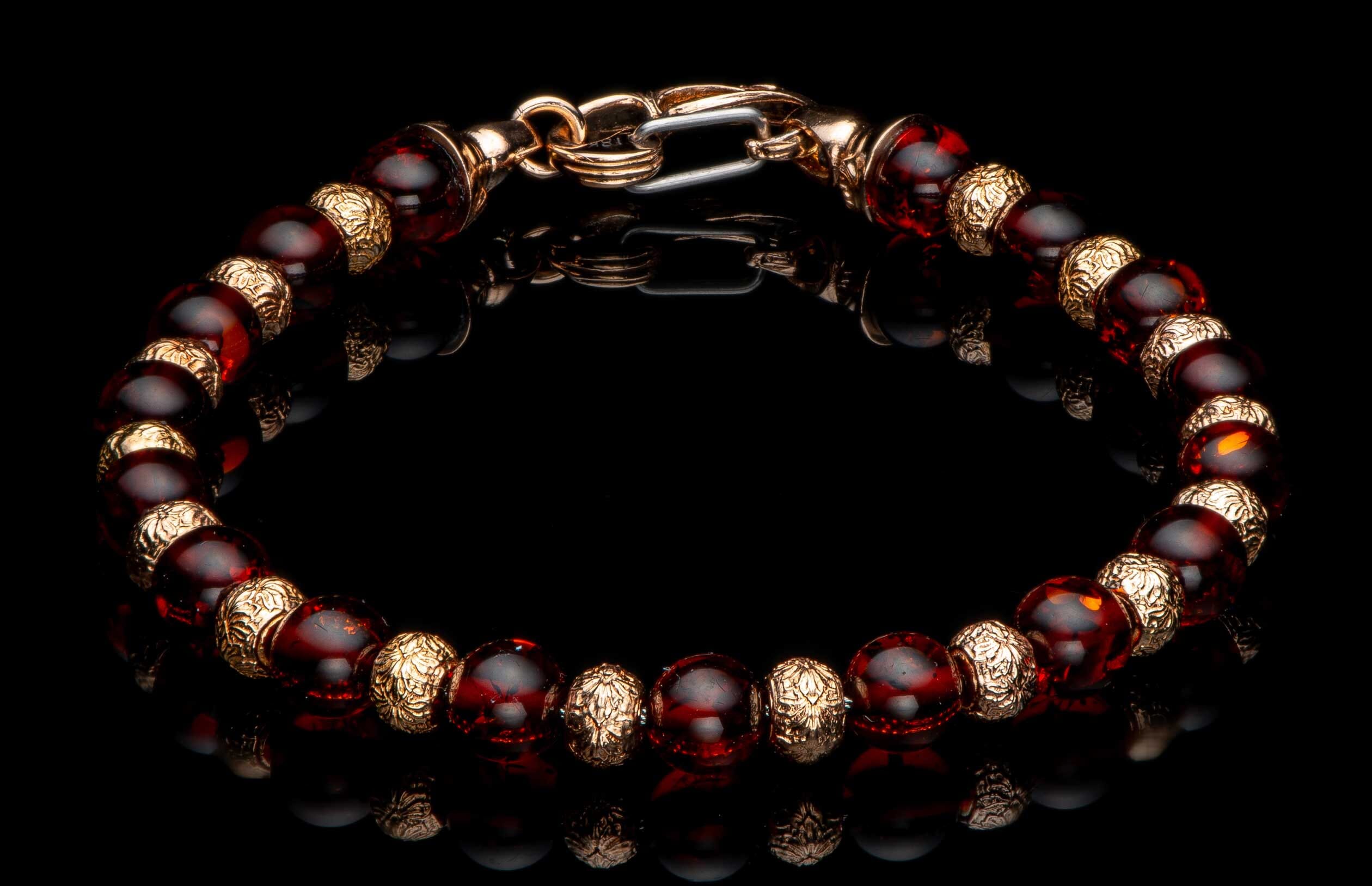 Beaded bracelet with reddish-brown amber-like beads, intricately patterned gold-toned spacers, and a lobster clasp on a reflective black surface.