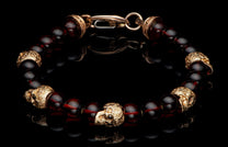Dark red amber bead bracelet with gold-colored skull charms and an ornate clasp, on a reflective black surface.
