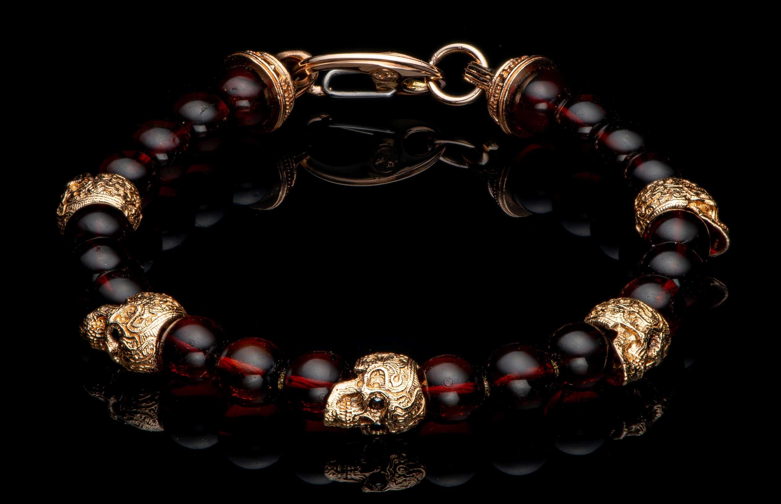 Dark red amber bead bracelet with gold-colored skull charms and an ornate clasp, on a reflective black surface.