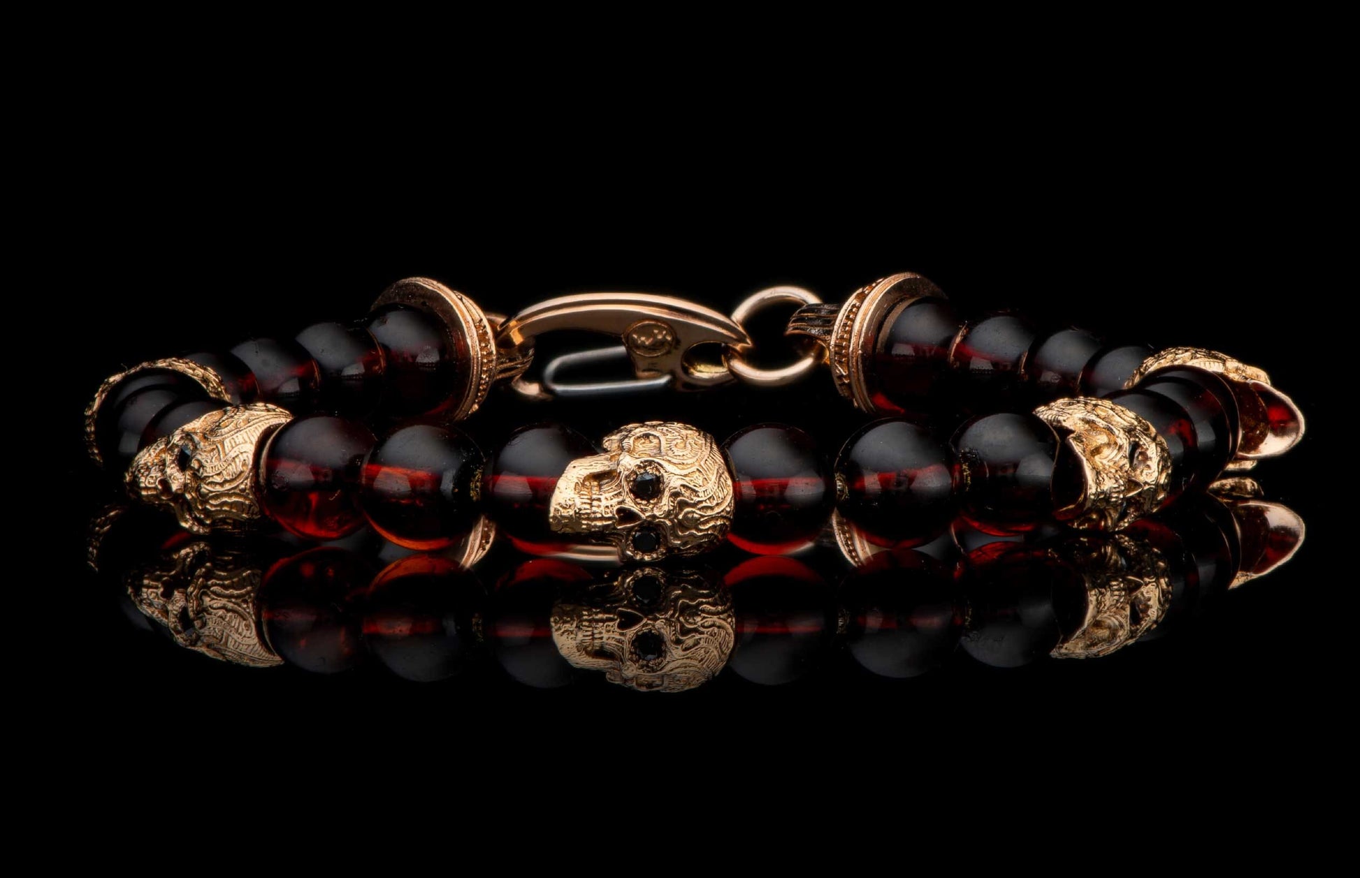 Dark amber beaded bracelet with gold skull charms and clasp, reflected on a black surface.
