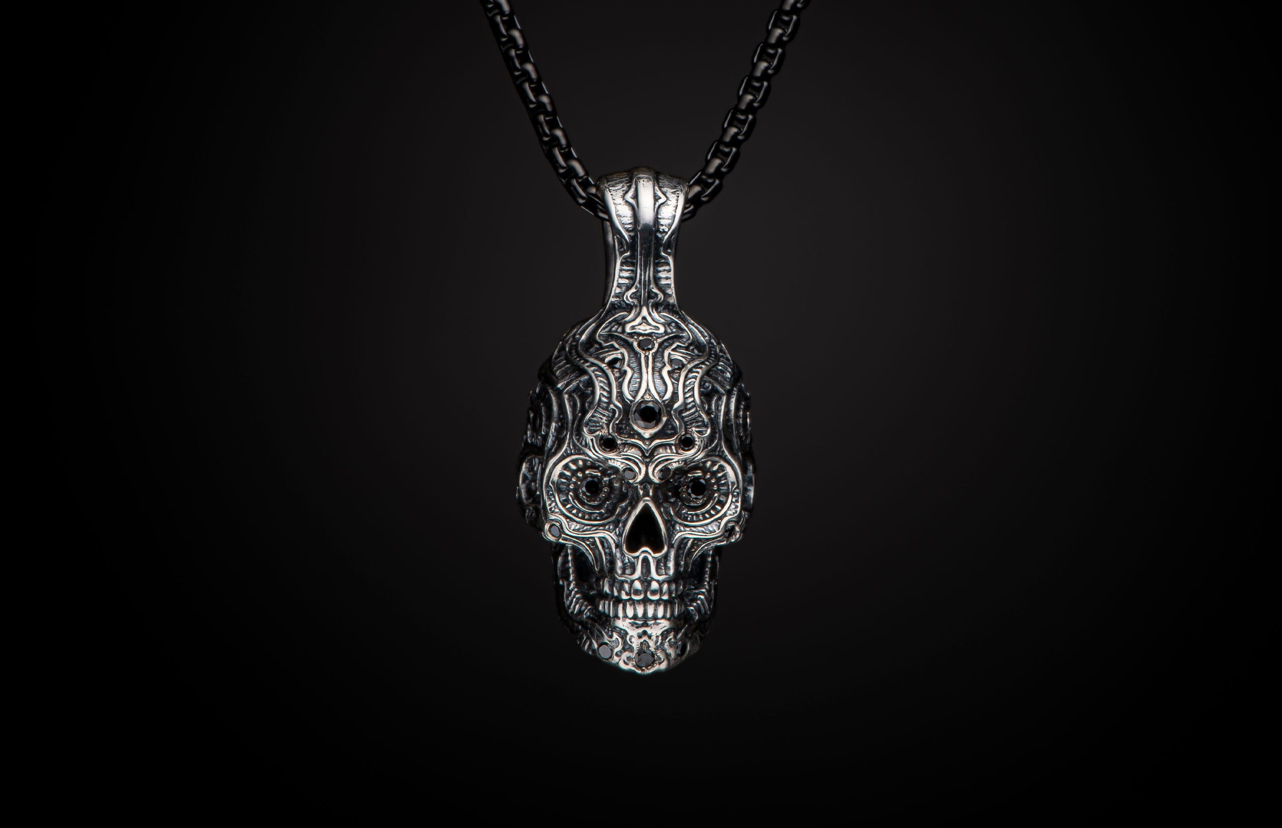 Front-facing silver skull pendant with intricate filigree patterns and black stone accents on a dark chain.