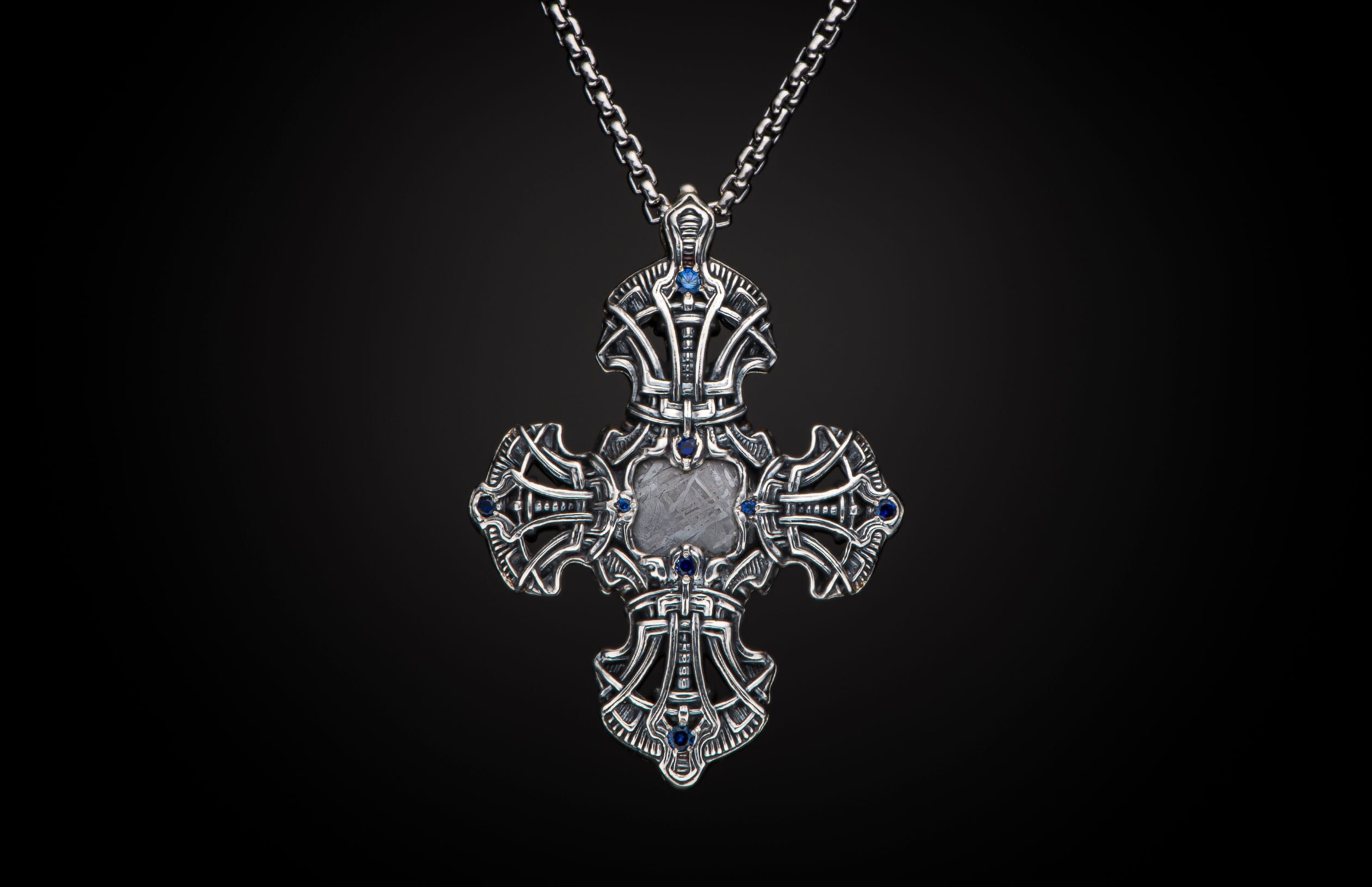Intricately designed silver cross pendant featuring blue gemstones and a rectangular meteorite inlay with Widmanstätten pattern, on a silver chain.
