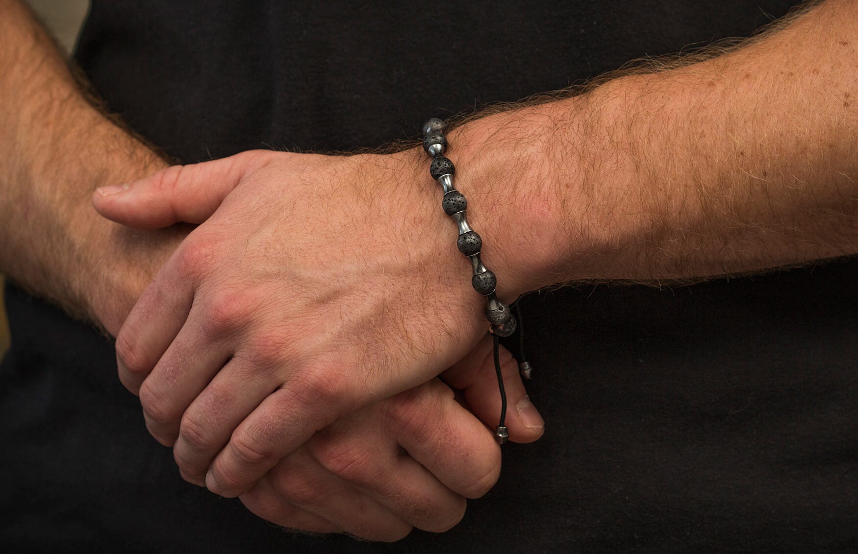 Black lava bead bracelet with silver-tone spacers and black cord, worn on a man's hairy arm.