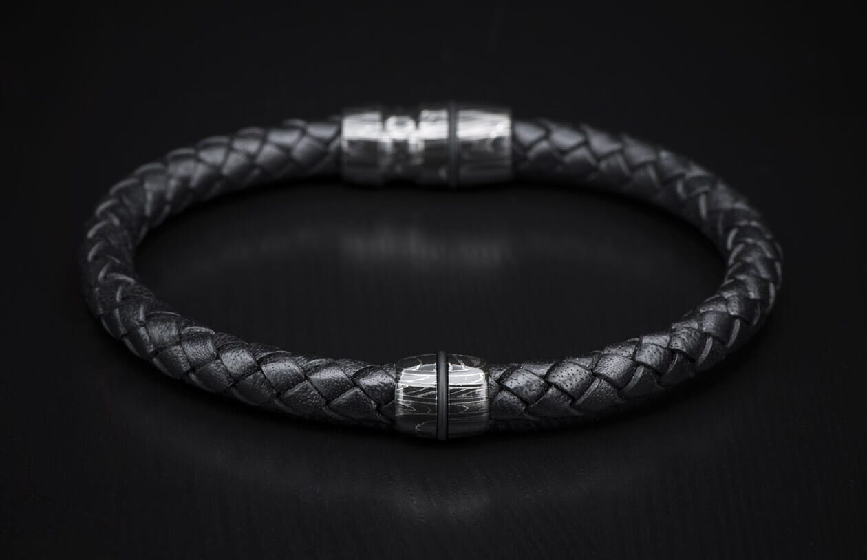 Black braided leather bracelet with a silver metal bead featuring a wavy pattern and a blurred magnetic clasp.