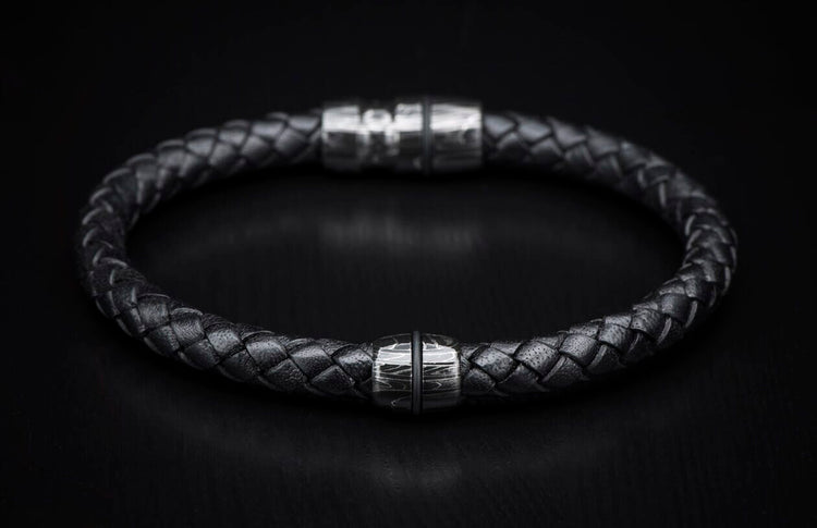 Black braided leather bracelet with a silver metal bead featuring a wavy pattern and a blurred magnetic clasp.