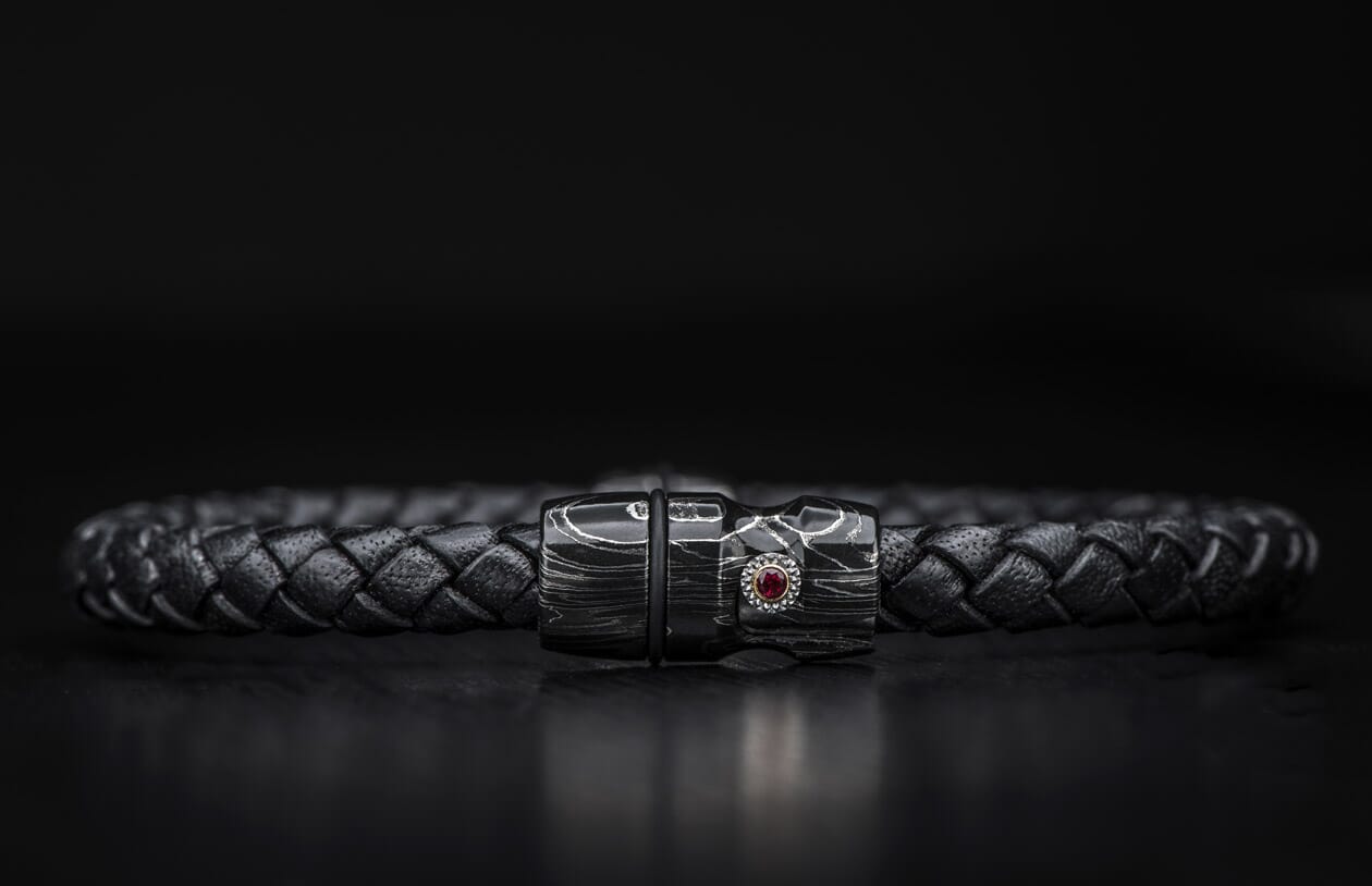 Black braided leather bracelet with a dark metallic clasp featuring a swirling pattern and a red gemstone.