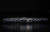 Black braided leather bracelet with a dark metallic clasp featuring a swirling pattern and a red gemstone.