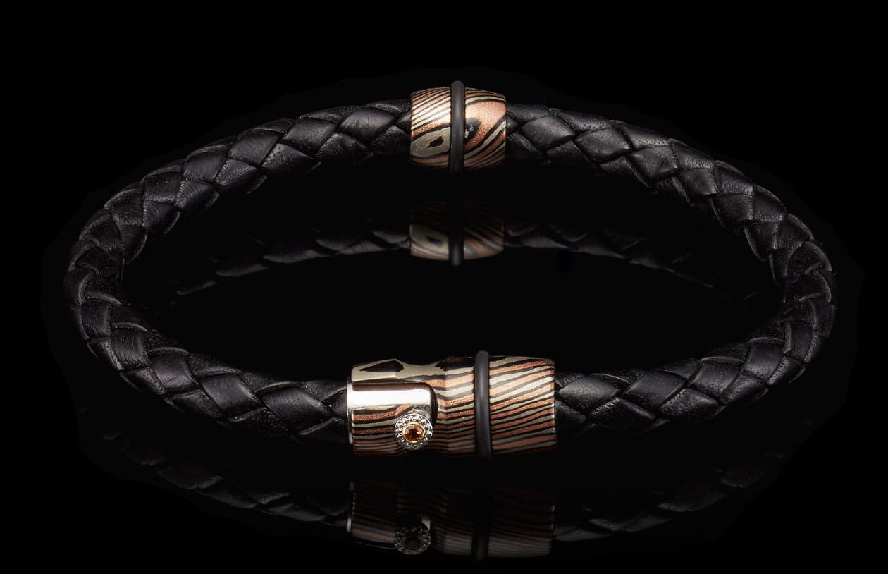 Black braided leather bracelet with two copper and silver patterned metal beads, one accented with an orange gemstone.
