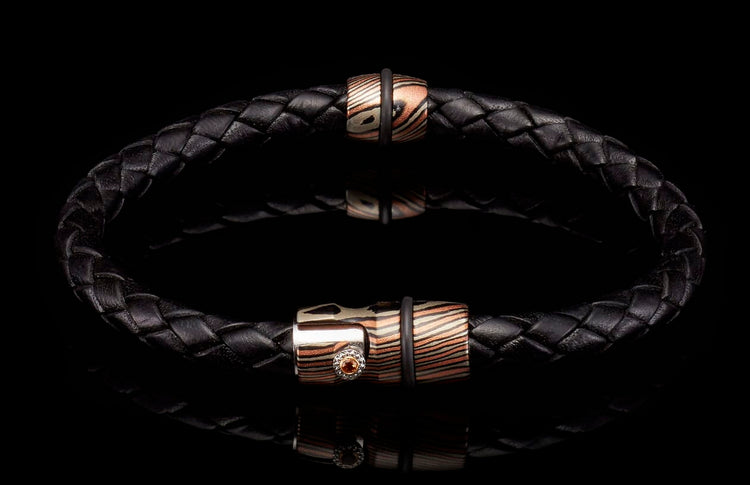 Black braided leather bracelet with two copper and silver patterned metal beads, one accented with an orange gemstone.