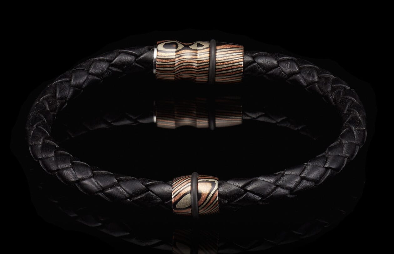 Black braided leather bracelet with two striped copper, silver, and black beads, each with a black rubber ring, on a reflective surface.
