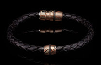 Black braided leather bracelet with two striped copper, silver, and black beads, each with a black rubber ring, on a reflective surface.