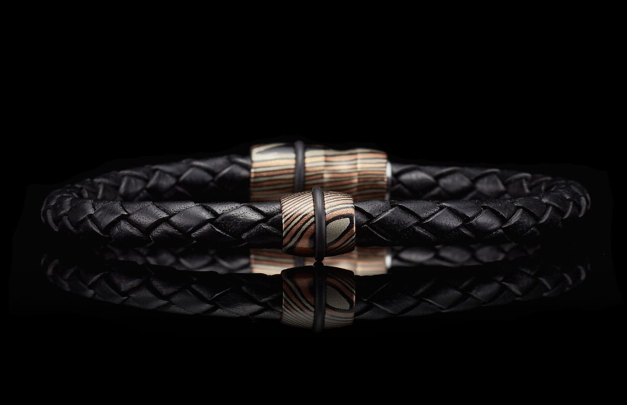 Black braided leather bracelet with two cylindrical copper and silver patterned beads, reflected on a dark surface.