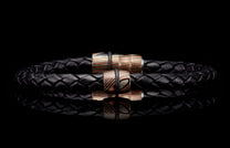 Black braided leather bracelet with two cylindrical copper and silver patterned beads, reflected on a dark surface.
