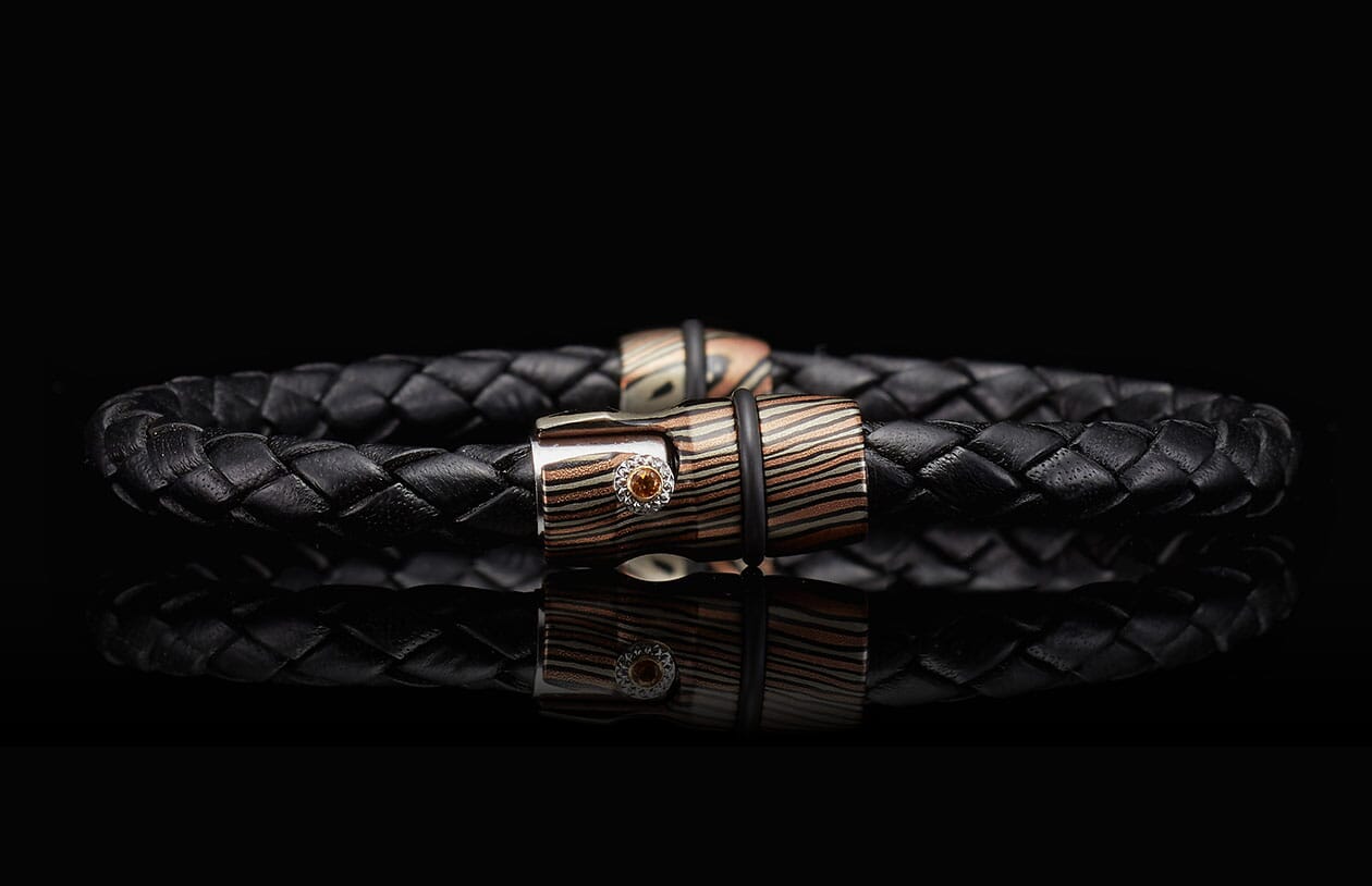 Black braided leather bracelet with a striped metallic clasp in bronze and silver tones, featuring an orange gemstone.