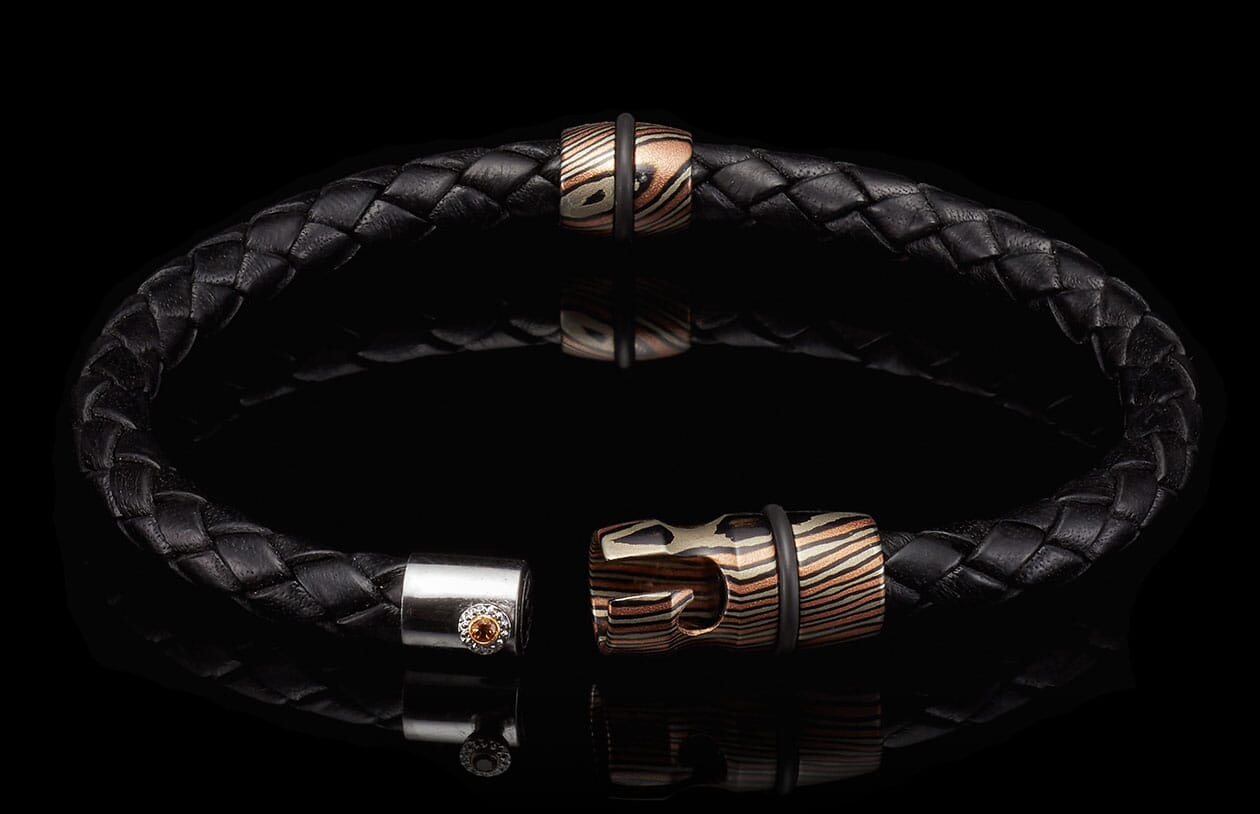 Black braided leather bracelet with mokume-gane patterned beads, black rubber rings, and an open clasp featuring an orange gemstone.