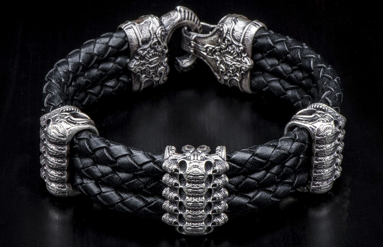 Black braided leather bracelet with ornate antique silver lion head clasp and stacked skull centerpiece.