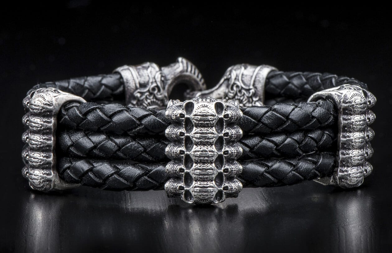 Black braided leather bracelet with silver skull-like center bead and ornate silver end caps, featuring a hook clasp.