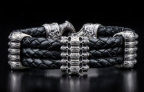 Black braided leather bracelet with silver skull-like center bead and ornate silver end caps, featuring a hook clasp.
