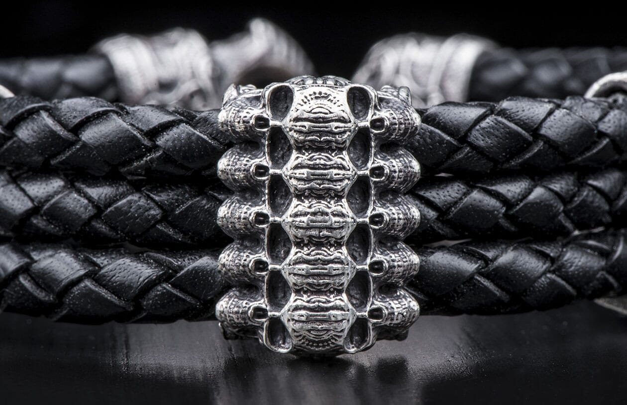 Black braided leather bracelet with an intricate silver-toned metal centerpiece featuring a stacked skull motif, resting on a dark surface.