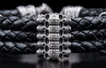 Black braided leather bracelet with an intricate silver-toned metal centerpiece featuring a stacked skull motif, resting on a dark surface.