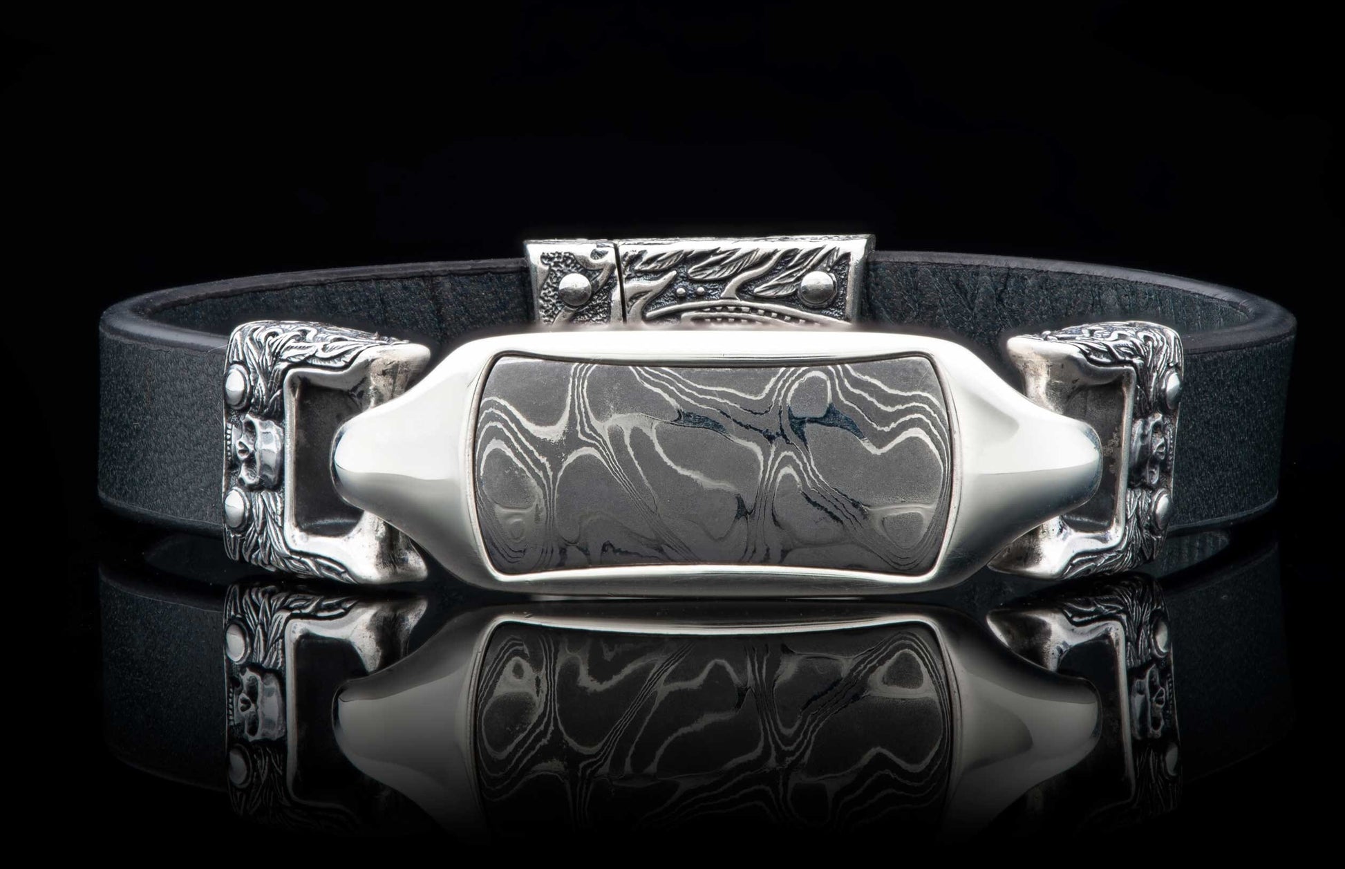 Black leather bracelet featuring a Damascus steel inlay framed by ornate silver-toned metalwork with skull details.