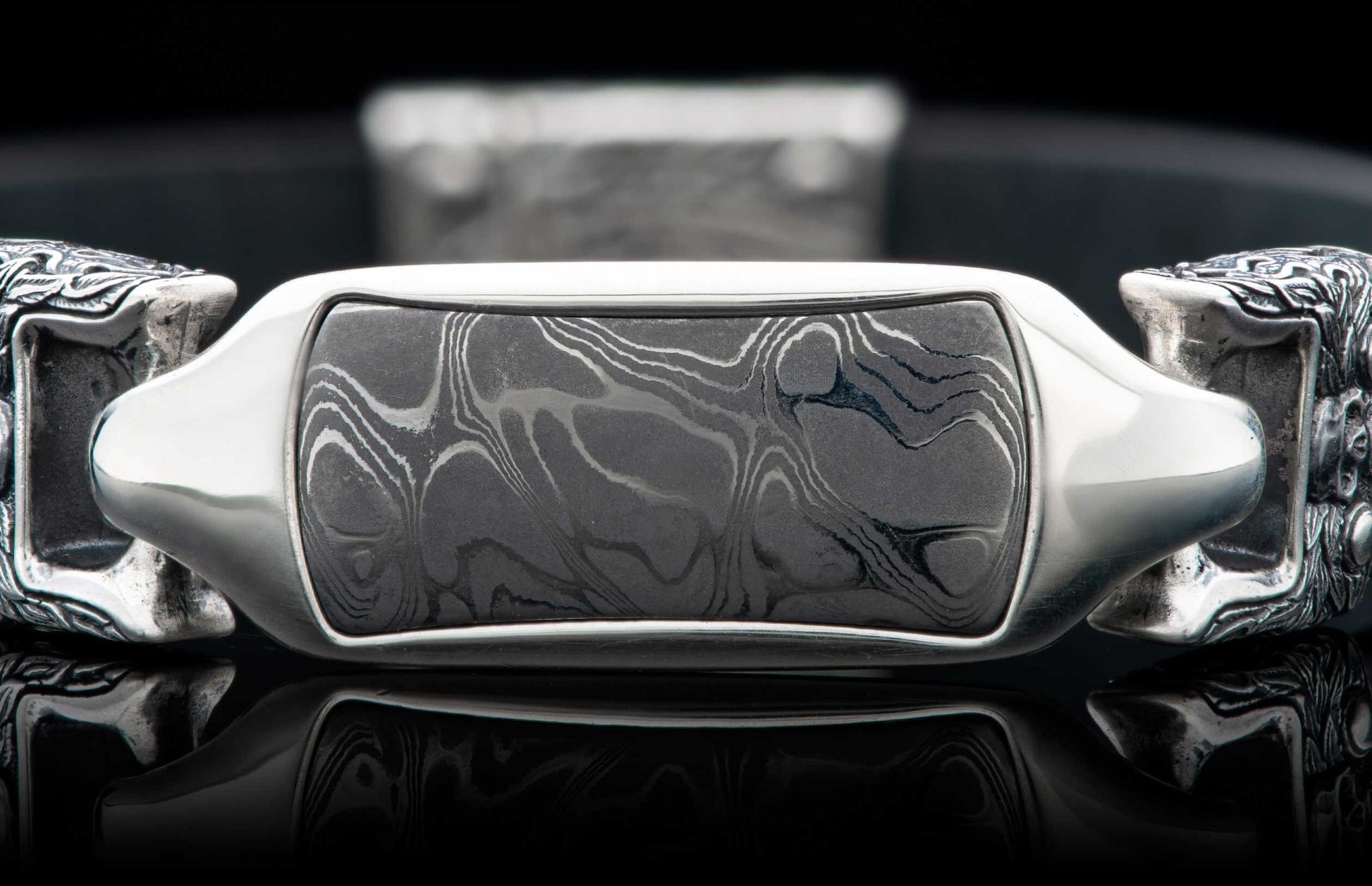 Damascus steel bracelet with a wavy pattern inlay, polished silver frame, ornate end caps, and reflective surface.