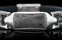 Damascus steel bracelet with a wavy pattern inlay, polished silver frame, ornate end caps, and reflective surface.