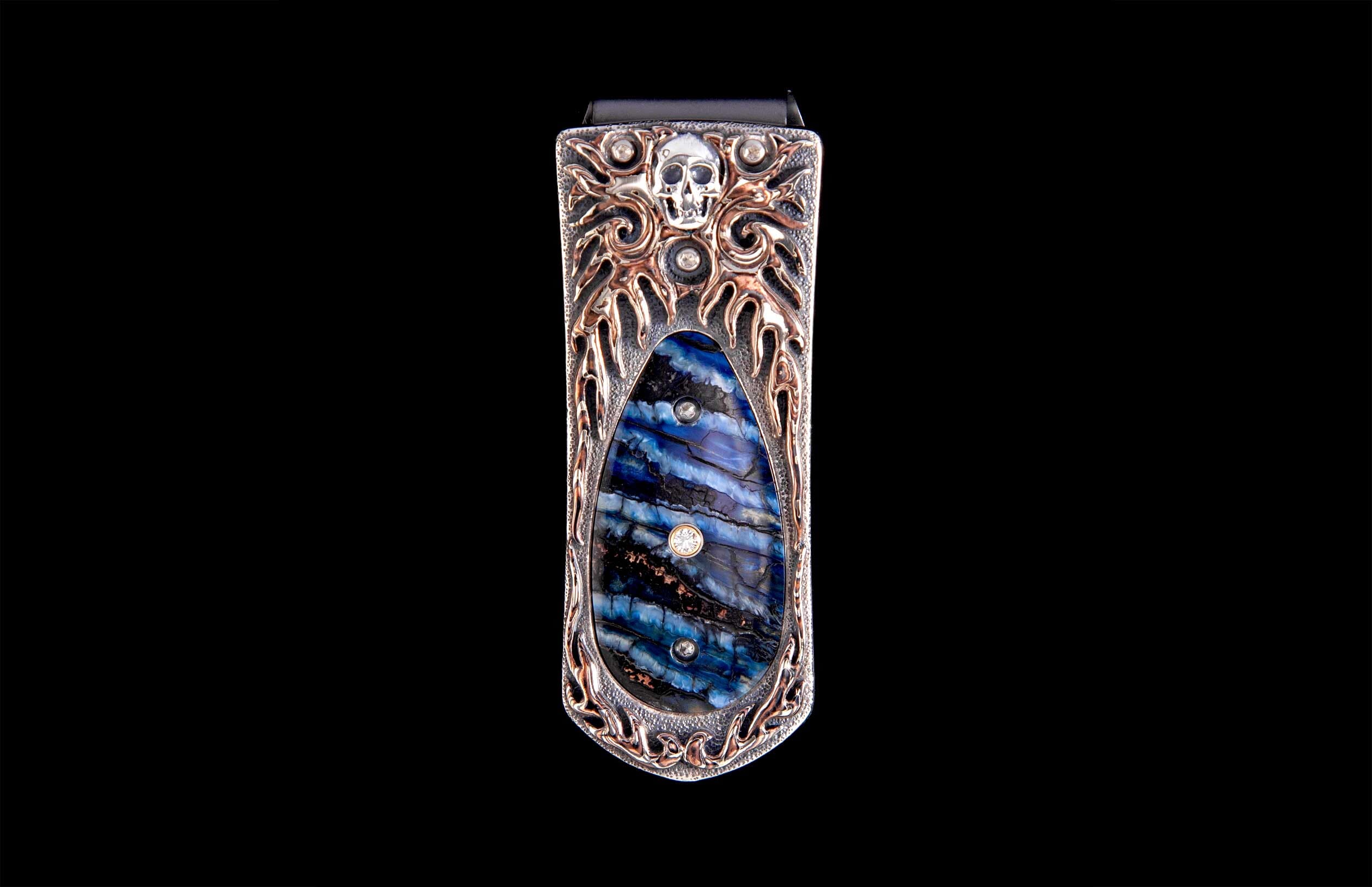 Money clip with a silver skull, ornate copper metalwork, and a blue and black striped inlay adorned with three diamonds.
