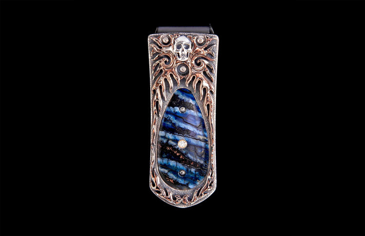 Money clip with a silver skull, ornate copper metalwork, and a blue and black striped inlay adorned with three diamonds.