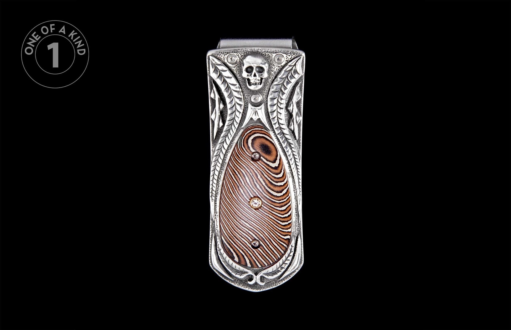 Silver money clip with an embossed skull, ornate carvings, and a brown Damascus patterned inlay with two gems.