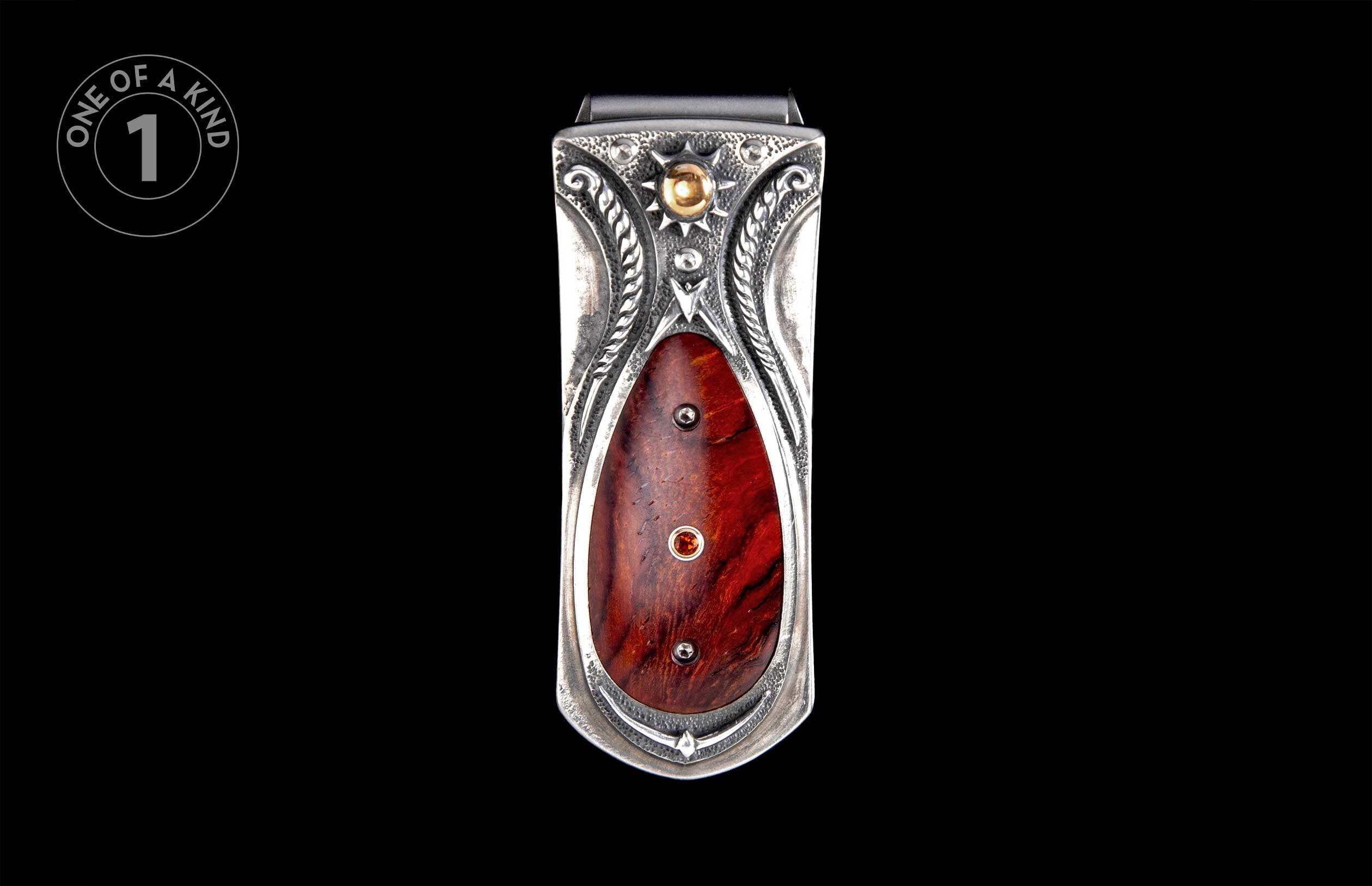 Ornate silver money clip featuring intricate engravings, a gold sunburst with a spherical accent, and a reddish-brown wood inlay with a red gemstone.