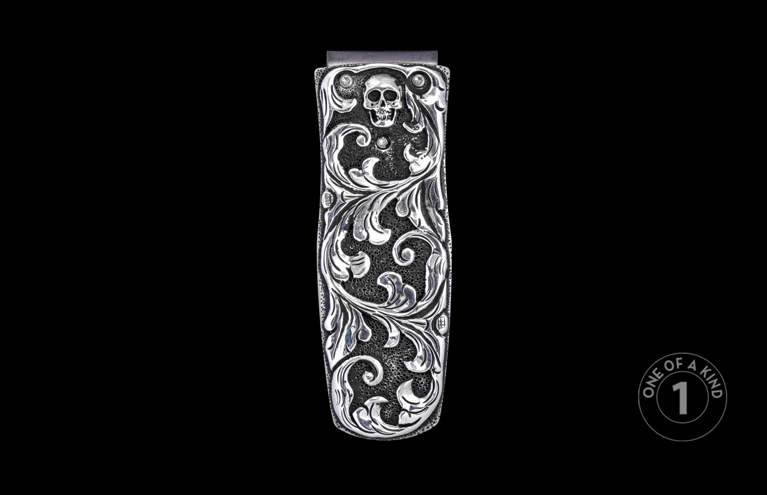 Silver-toned metal money clip with intricate scrollwork, skull design, and clear stone accents on a pebbled background.