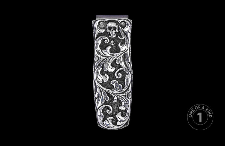 Silver-toned metal money clip with intricate scrollwork, skull design, and clear stone accents on a pebbled background.
